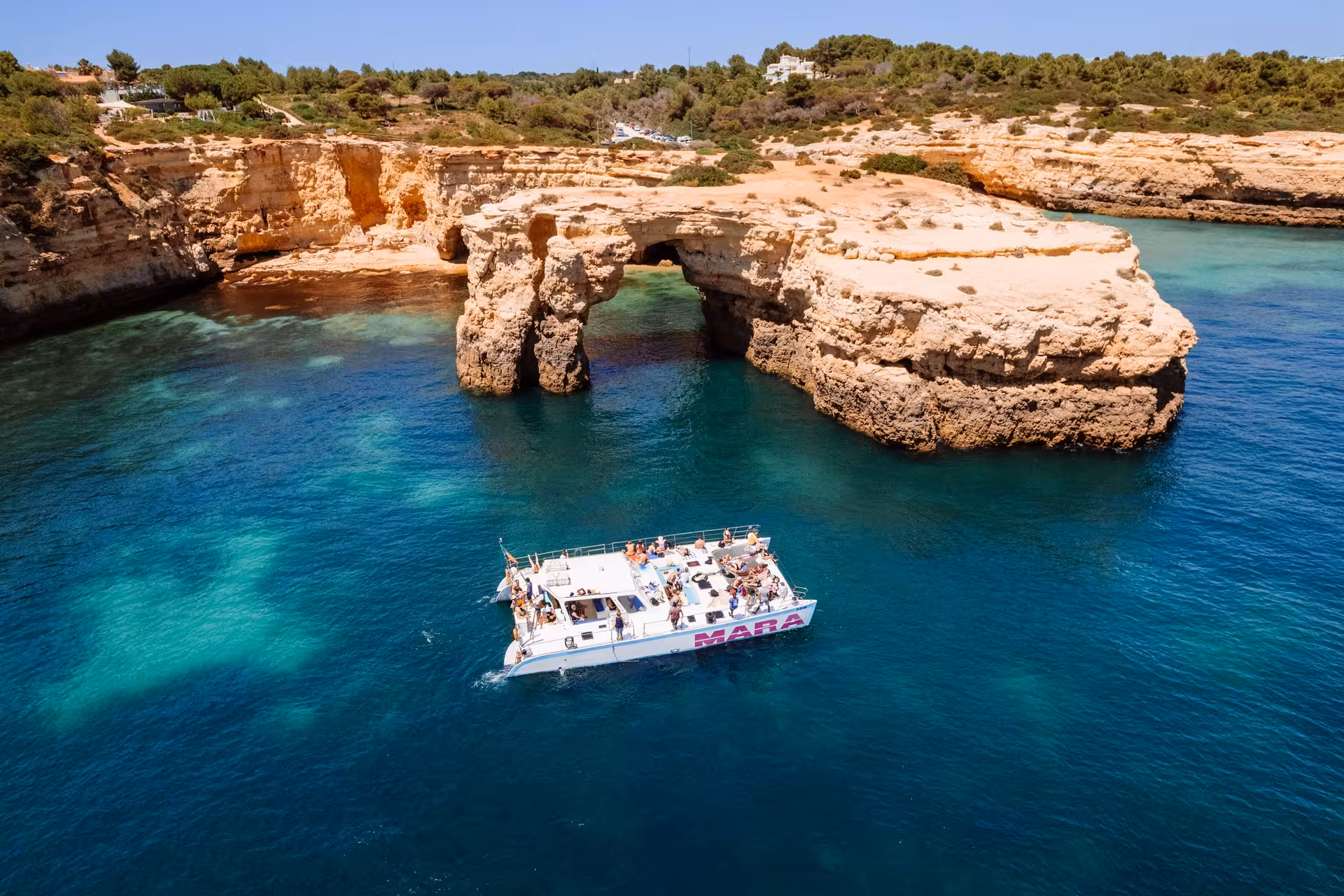 Catamaran MARA navigates turquoise waters near stunning coastal cliffs and natural arches, offering scenic Algarve views.