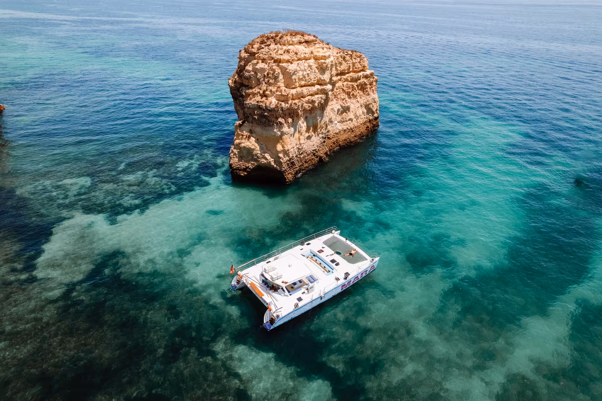 Catamaran MARA cruises near a stunning rock formation in clear turquoise waters, perfect for a beach BBQ adventure.