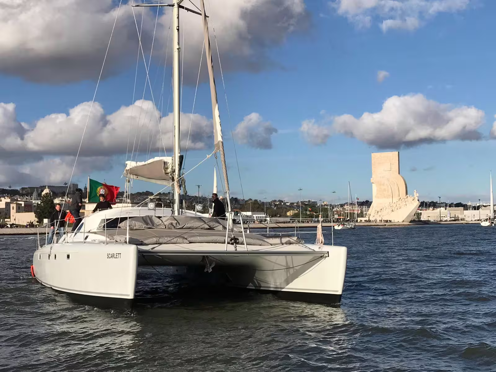 Catamaran Scarlett sailing past Lisbon's Discoveries Monument with Portuguese flag flying.