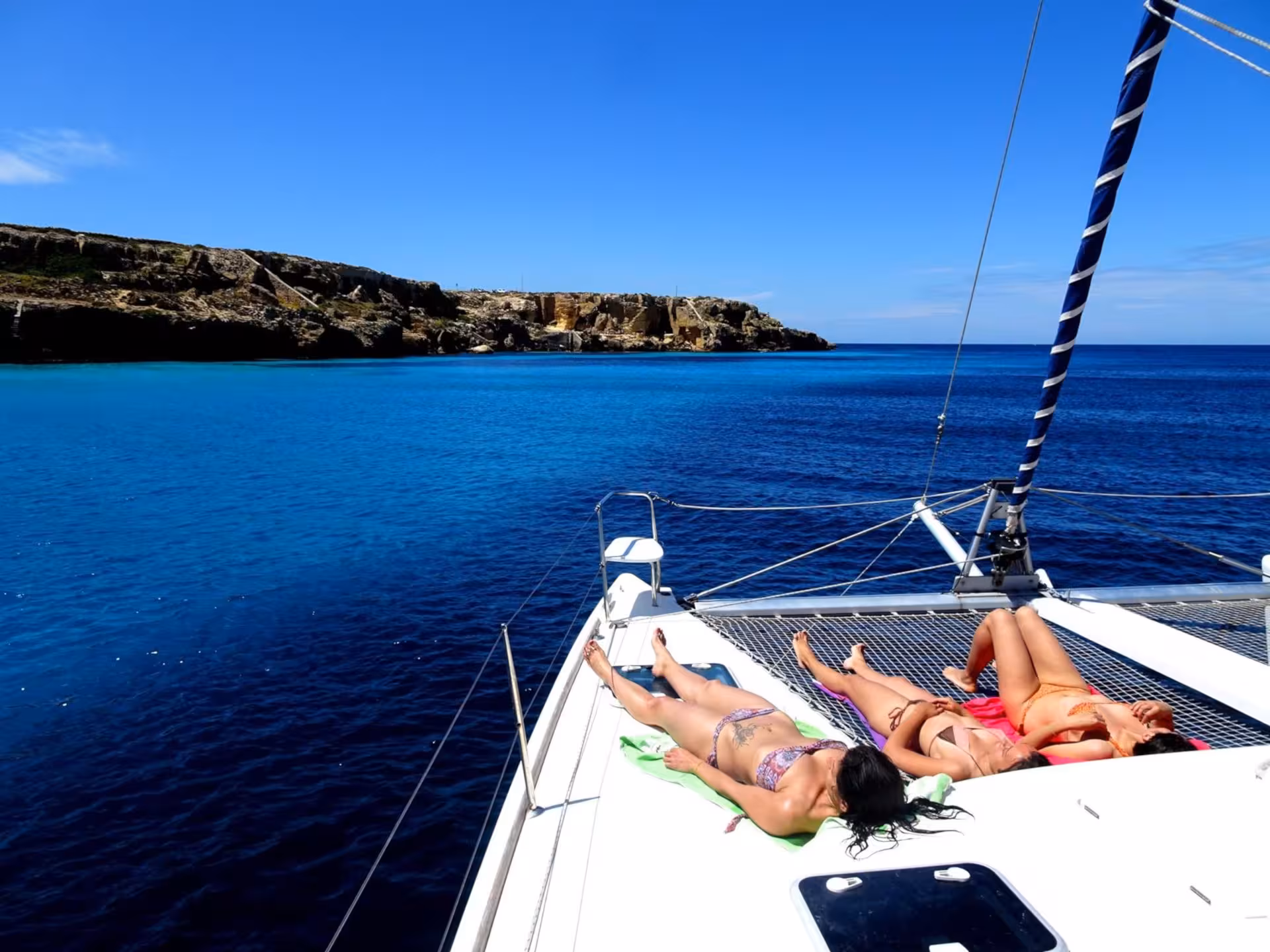 Sunbathers relaxing on a catamaran deck with stunning views of La Maddalena Archipelago's azure sea and cliffs.