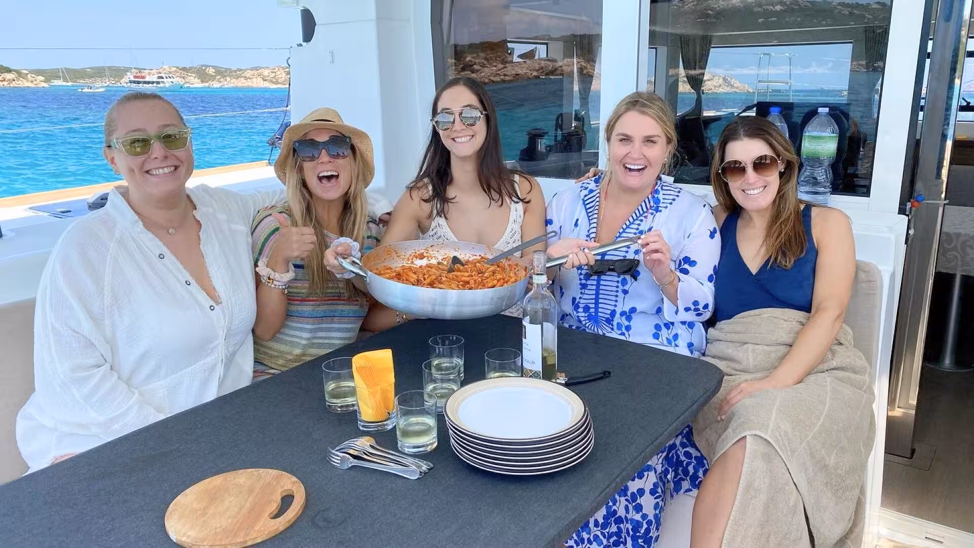 Group enjoying a pasta meal on a catamaran tour in La Maddalena archipelago, surrounded by turquoise waters.