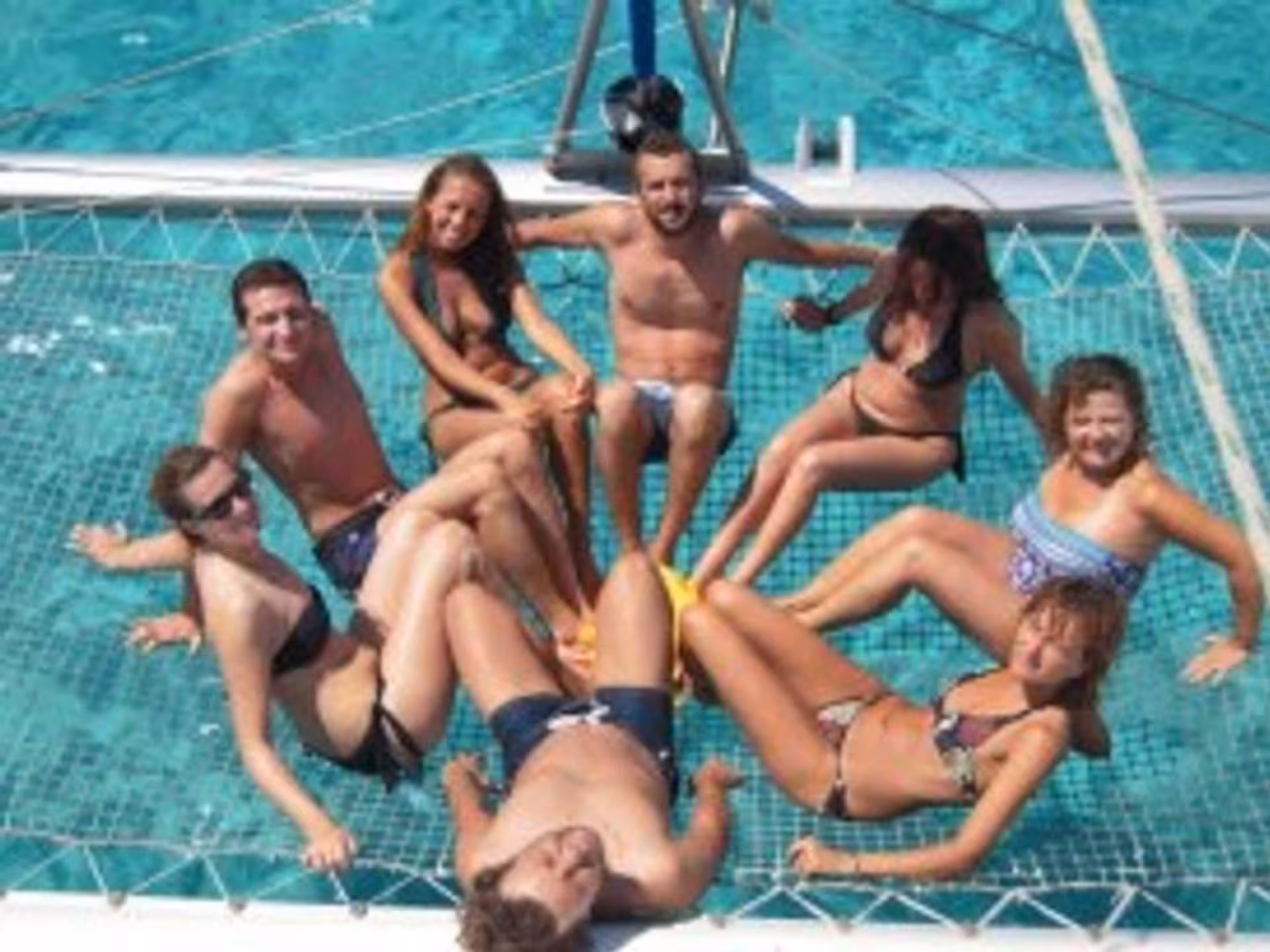 Group of friends relaxing on a catamaran net over turquoise waters in La Maddalena Archipelago.