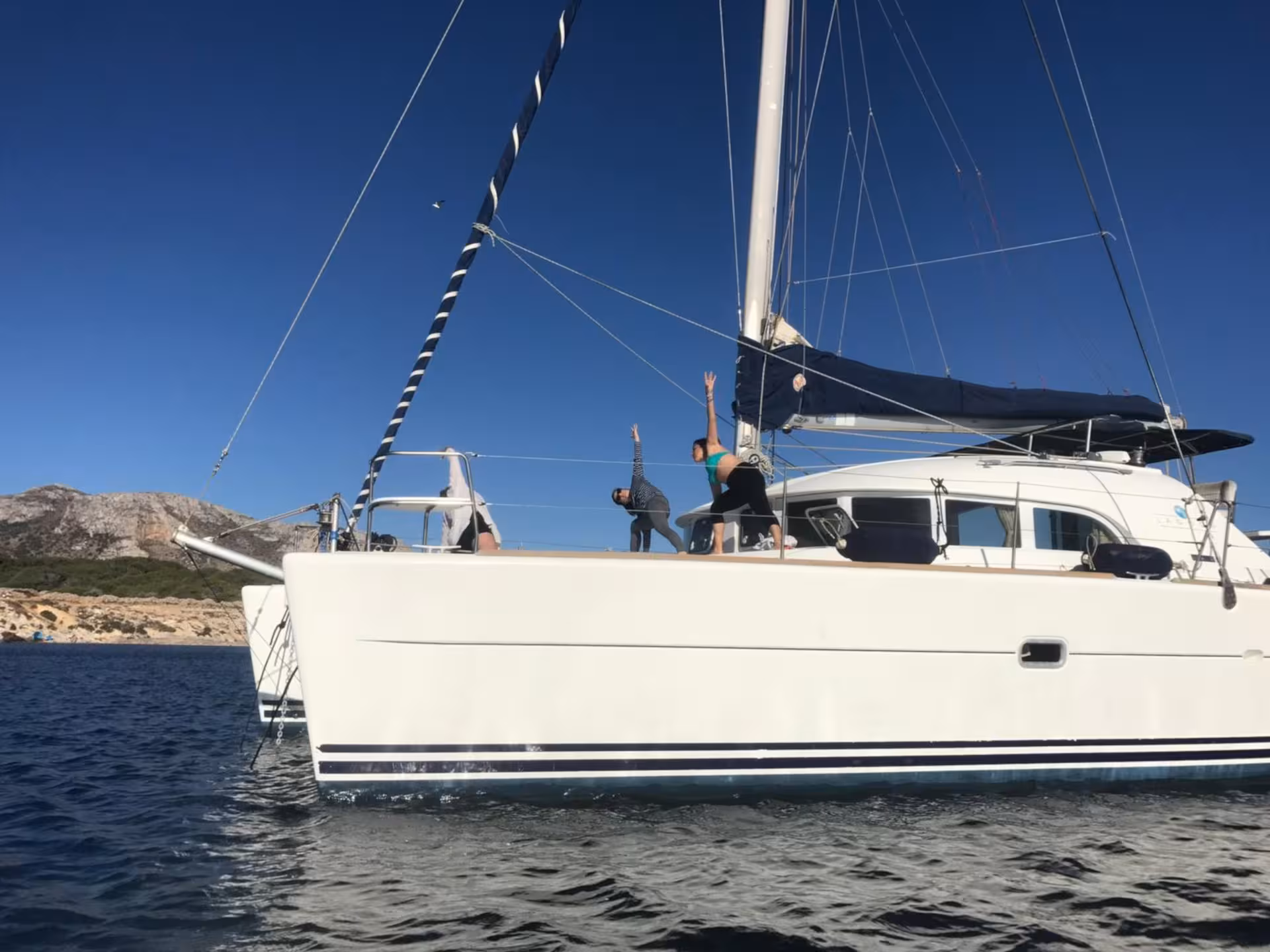 Catamaran Tenerè anchored near Cannigione with guests practicing yoga on deck against a mountainous backdrop.