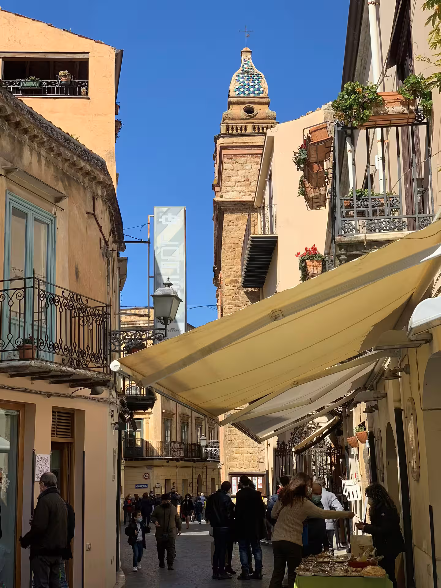Lively narrow street in Cefalù with tiled church tower, balconies and locals under awnings on a guided Palermo excursion