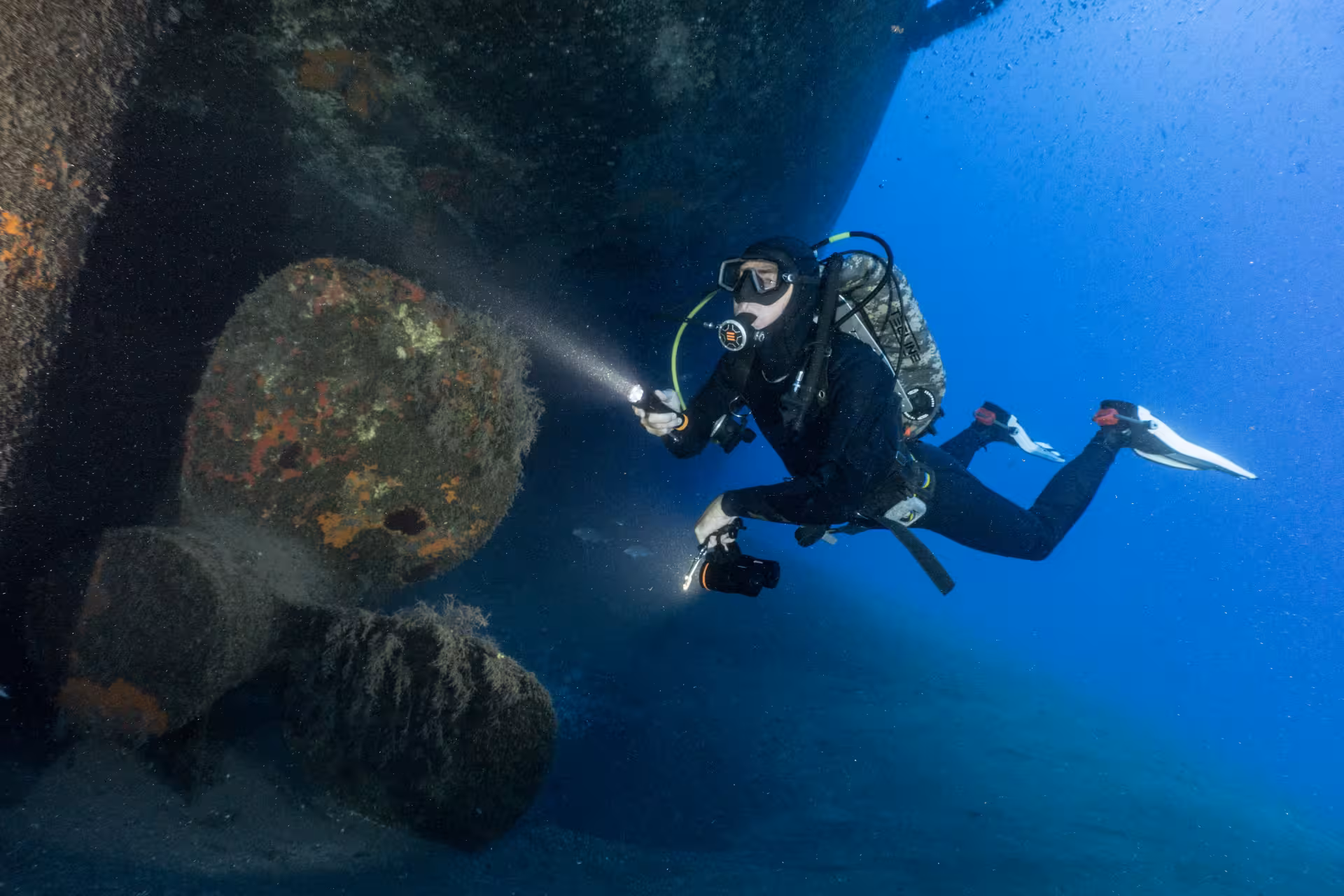 Diver explores underwater shipwreck with flashlight, highlighting vibrant marine life, ideal for certified diving tours.