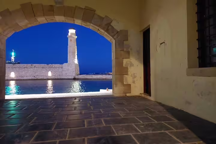 View of the illuminated Chania lighthouse through an archway at night, highlighting the charm of a private West Crete tour.