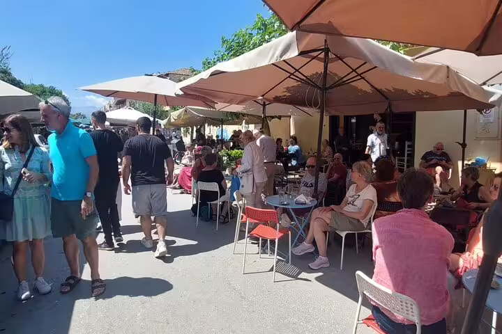 Visitors enjoying outdoor cafes and market atmosphere in Chania's Old Town, shaded by large umbrellas on a sunny day.