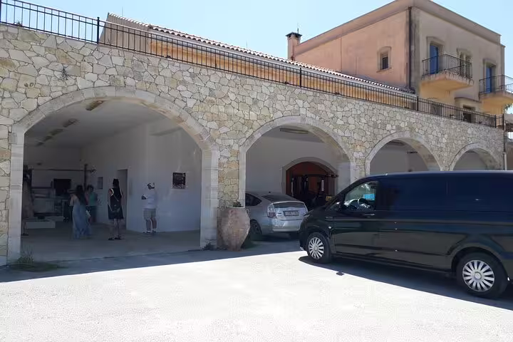 Exterior view of a Chania olive oil mill with stone arches, parked vehicles, and visitors entering the facility.