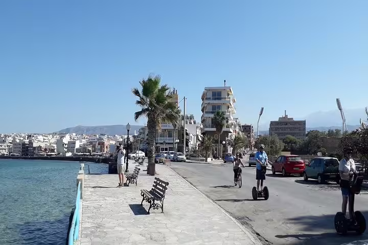 Coastal promenade in Chania, Crete, featuring tourists on Segways, scenic views, and Mediterranean architecture under a clear blue sky.