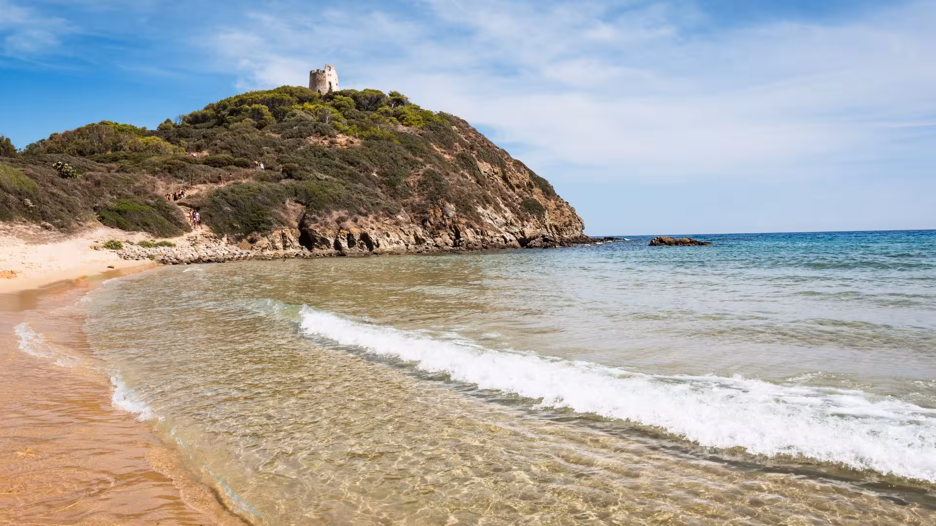 Stunning view of Chia beach with turquoise waters and a historic tower on a lush hillside, perfect for a dinghy tour.