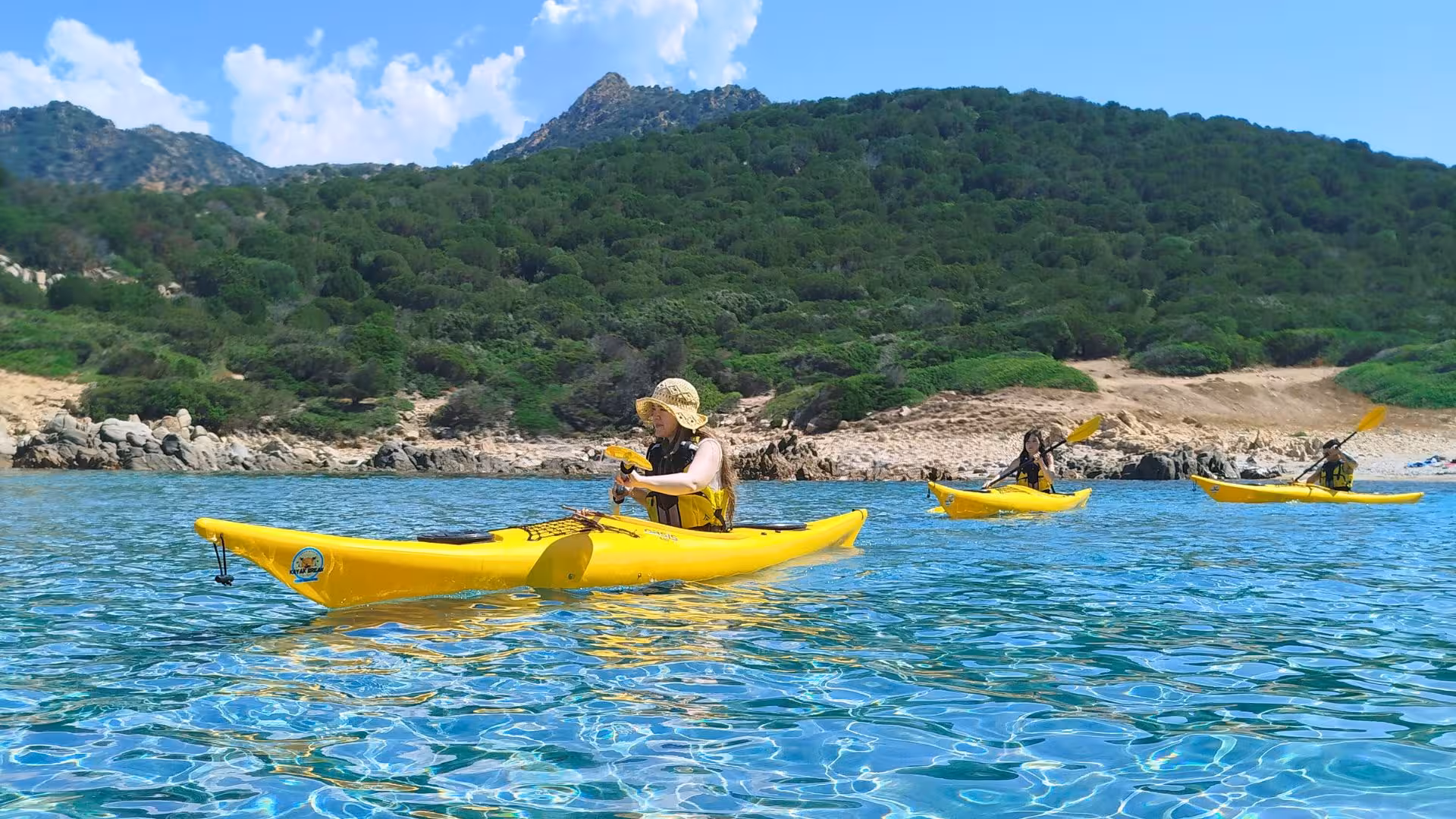 Kayakers paddle in vibrant yellow kayaks on clear turquoise waters near Tuerredda, surrounded by lush green hills.