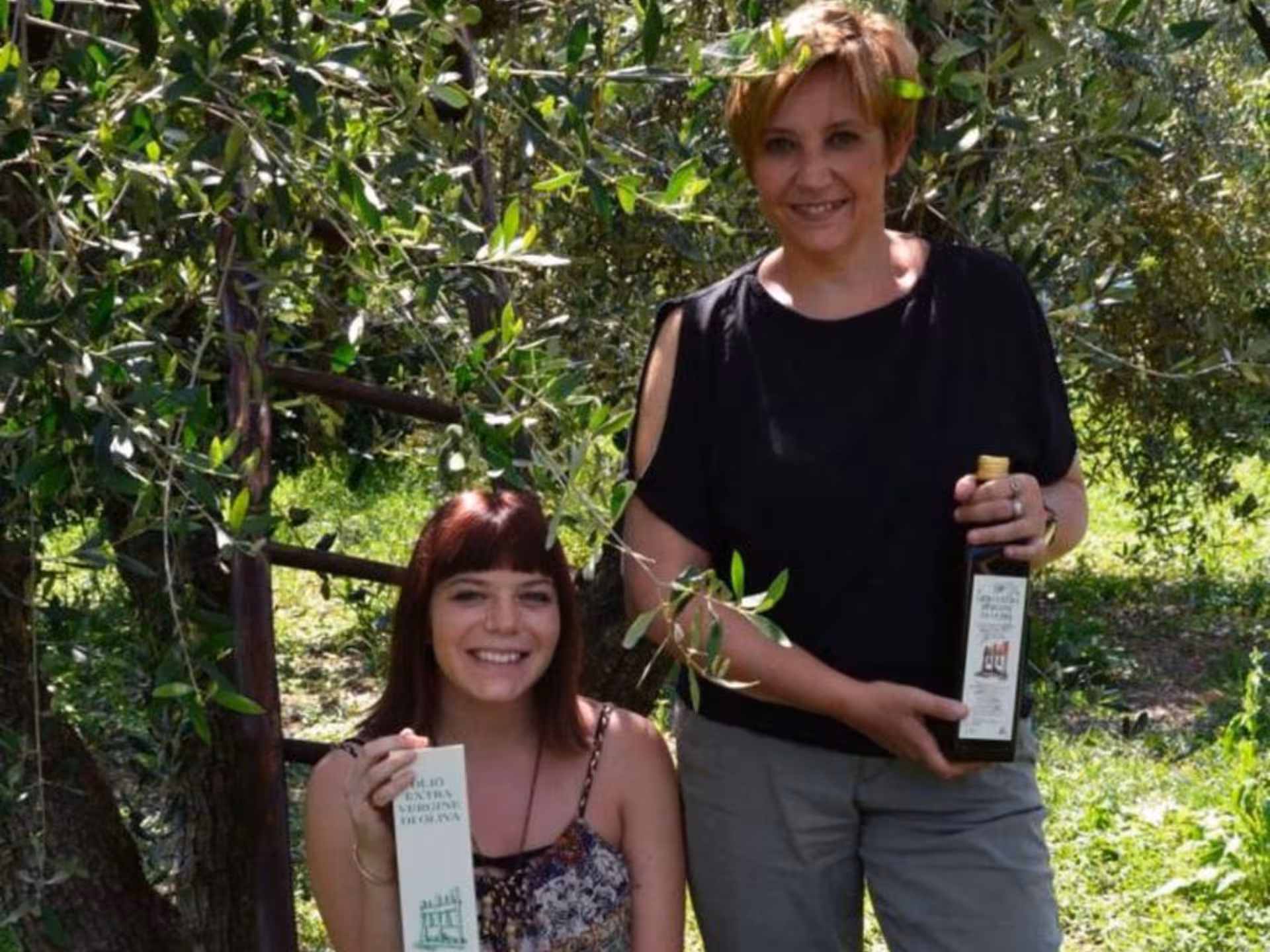 Two smiling women showcasing premium olive oil products during a tasting tour in a Chianciano olive grove.