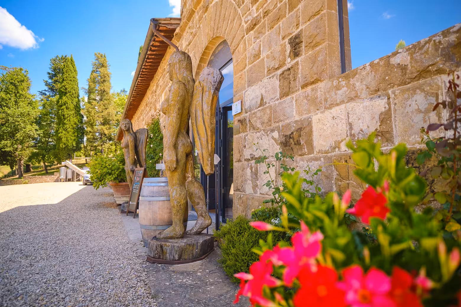 Rustic stone building with wooden sculptures and vibrant flowers at a Chianti vineyard entrance, Tuscany tour.