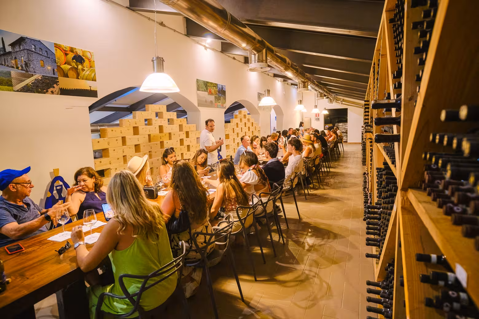 Large group of tourists at a long table in Chianti wine cellar, engaging in a guided wine tasting session.