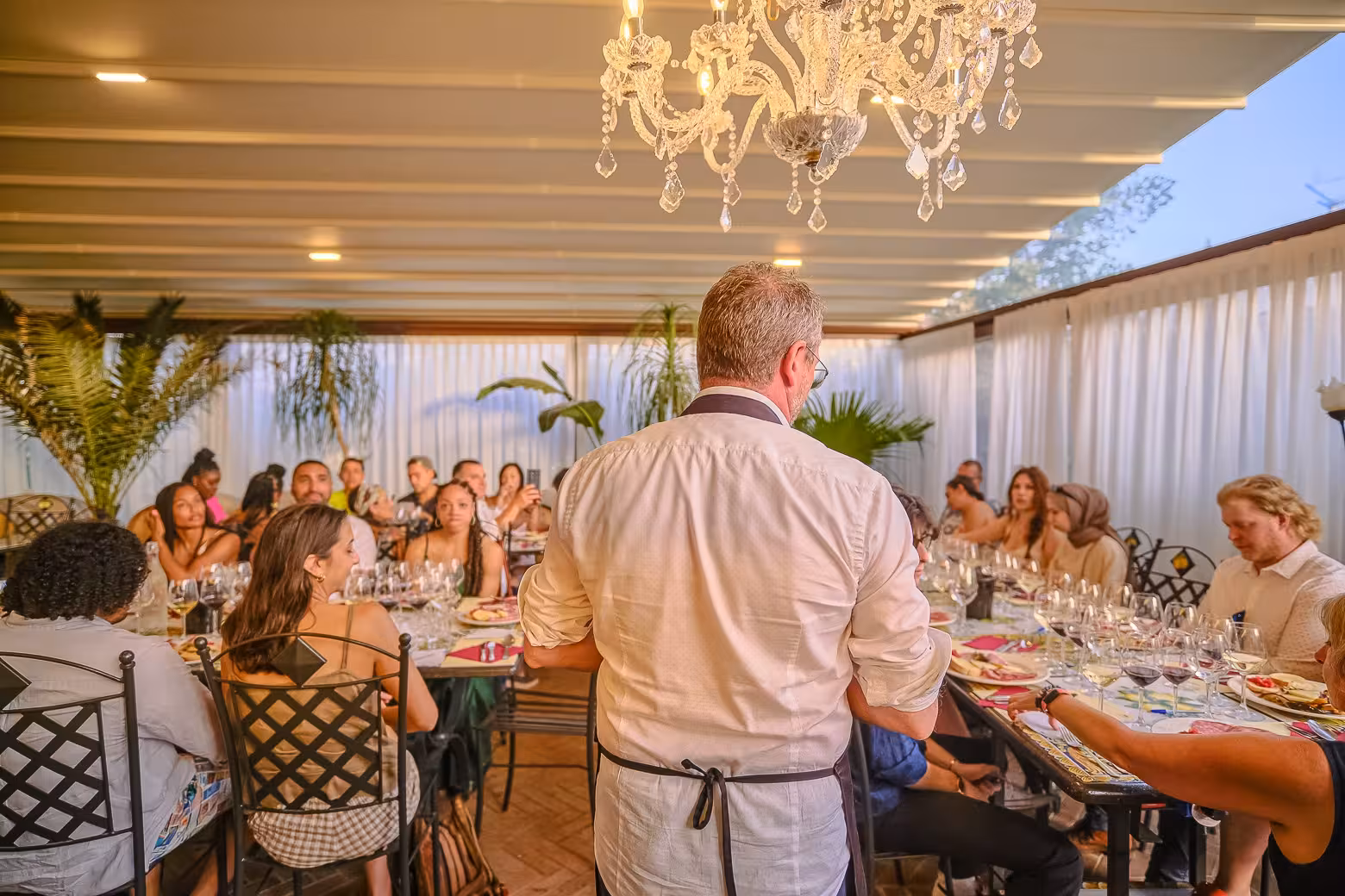 Winery host speaking to guests at an elegant Chianti dinner under a chandelier, surrounded by wine glasses.