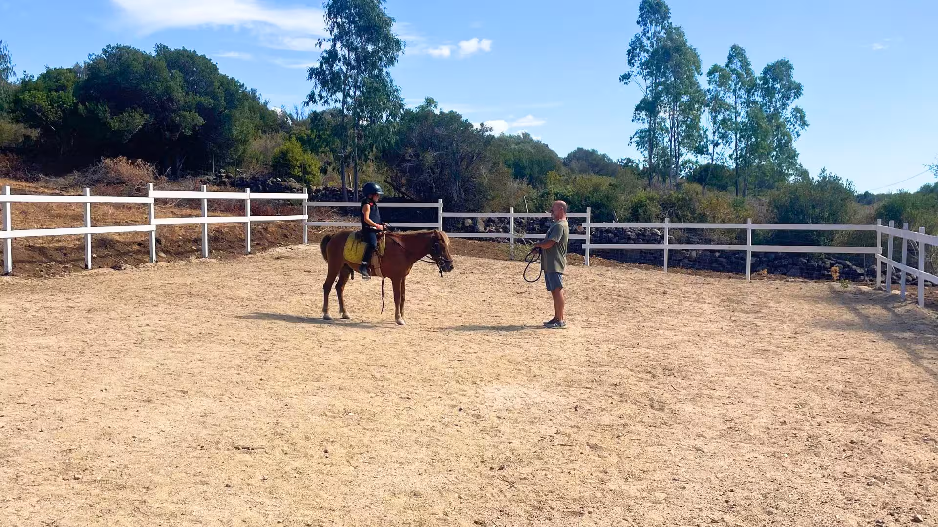 Child with helmet learning to ride a horse in a sunny Dorgali arena guided by an instructor.