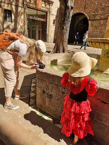 Child in a vibrant flamenco dress and sun hat enjoys a sunny day by a fountain in Barcelona during a family tour.
