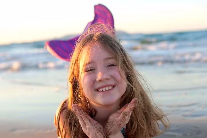 Smiling child with a purple mermaid tail poses happily on a sandy beach during sunset in Hersonissos.