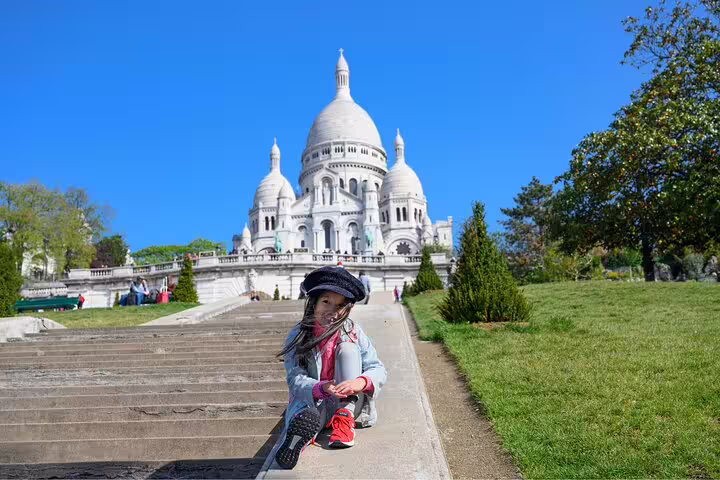 Child sitting on steps leading to Sacré-Cœur Basilica in Montmartre, Paris, under a clear blue sky.