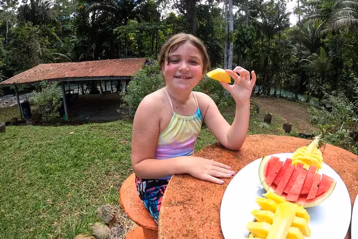 Child enjoying fresh tropical fruits at a scenic riverside spot near the Celeste River, amidst lush greenery.