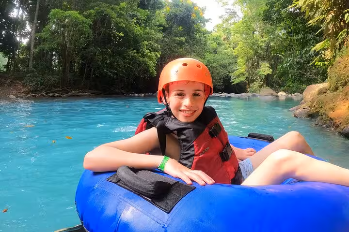 Smiling child in safety gear tubing on the vibrant turquoise waters of Celeste River, surrounded by lush rainforest.