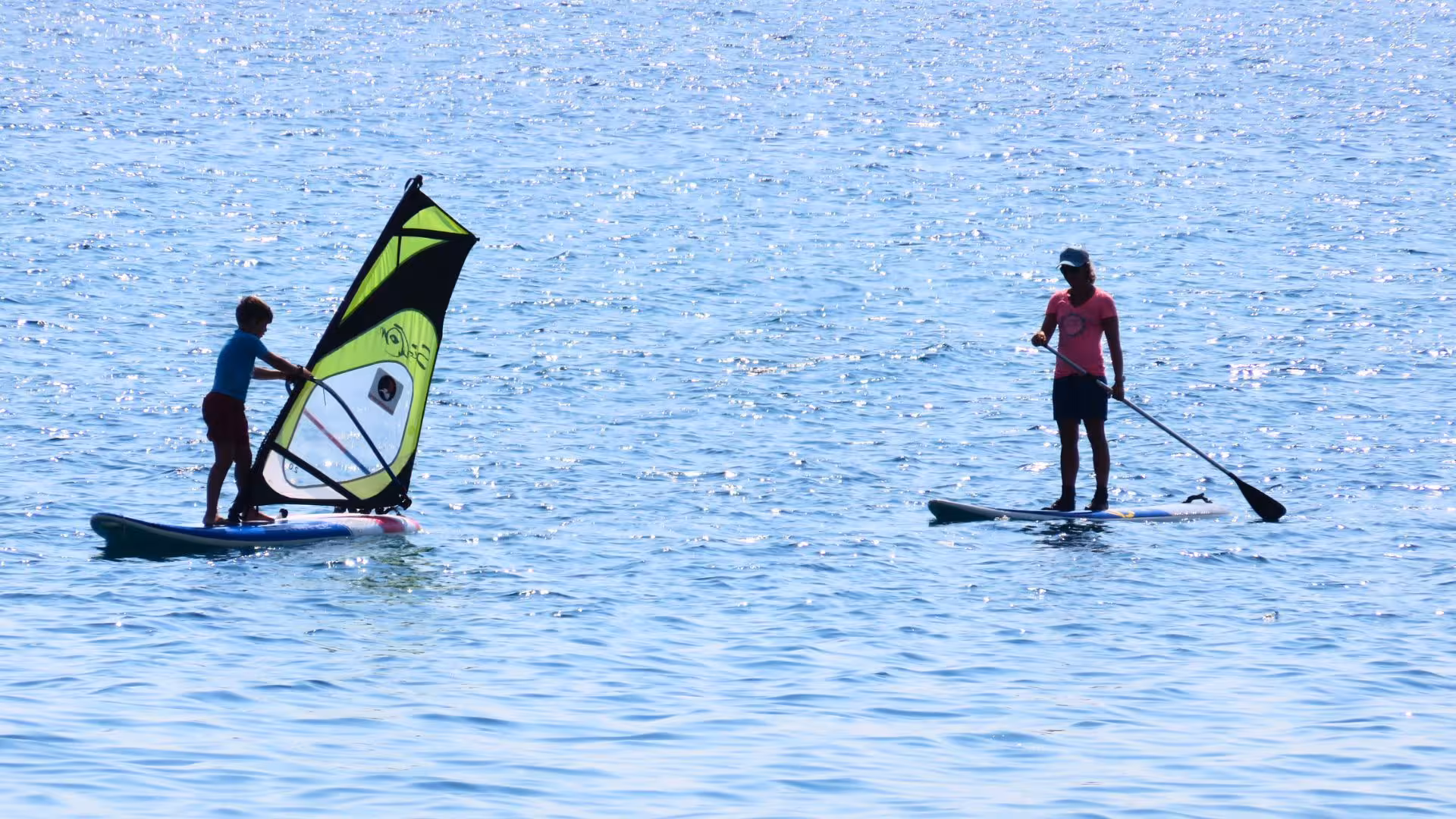 Child learning windsurfing in Bari Sardo's scenic waters, accompanied by an instructor during a basic course session.