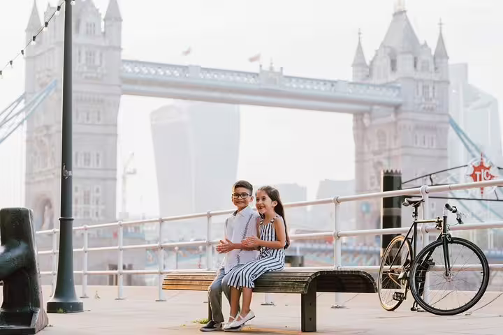 Two children smiling on a bench with Tower Bridge in the background during a London travel photographer tour.