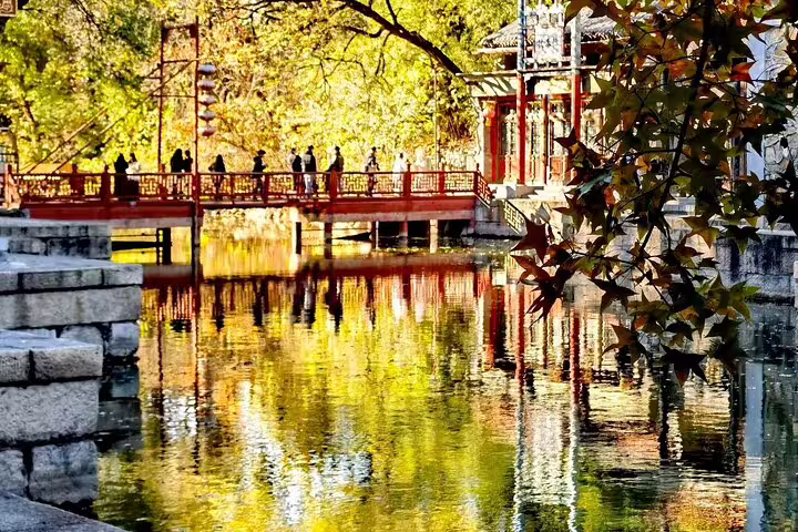 Visitors stroll across a vibrant red bridge reflecting autumn colors in a tranquil Chinese garden setting.