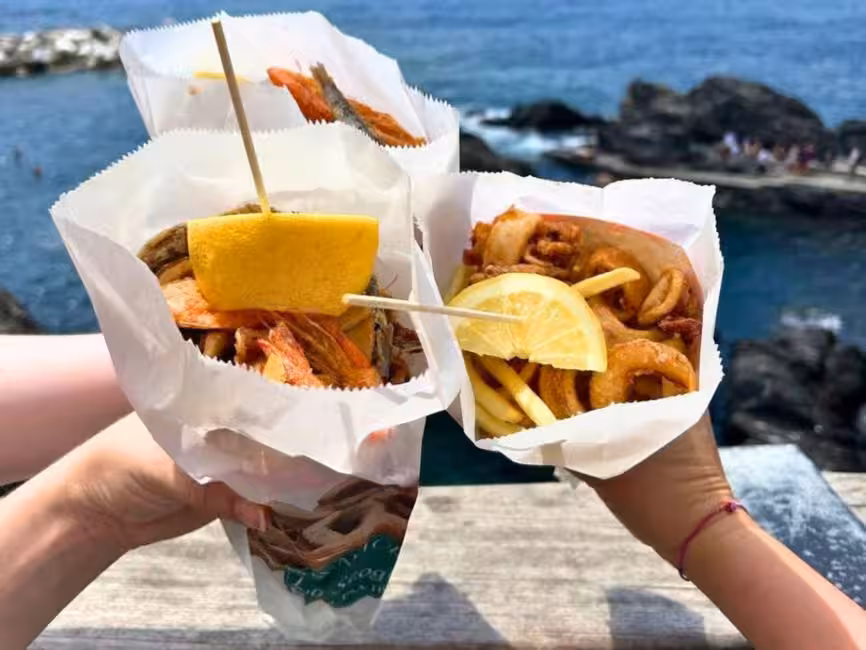 Delicious seafood cones with lemon slices held by tourists overlooking the rocky coastline of Cinque Terre.