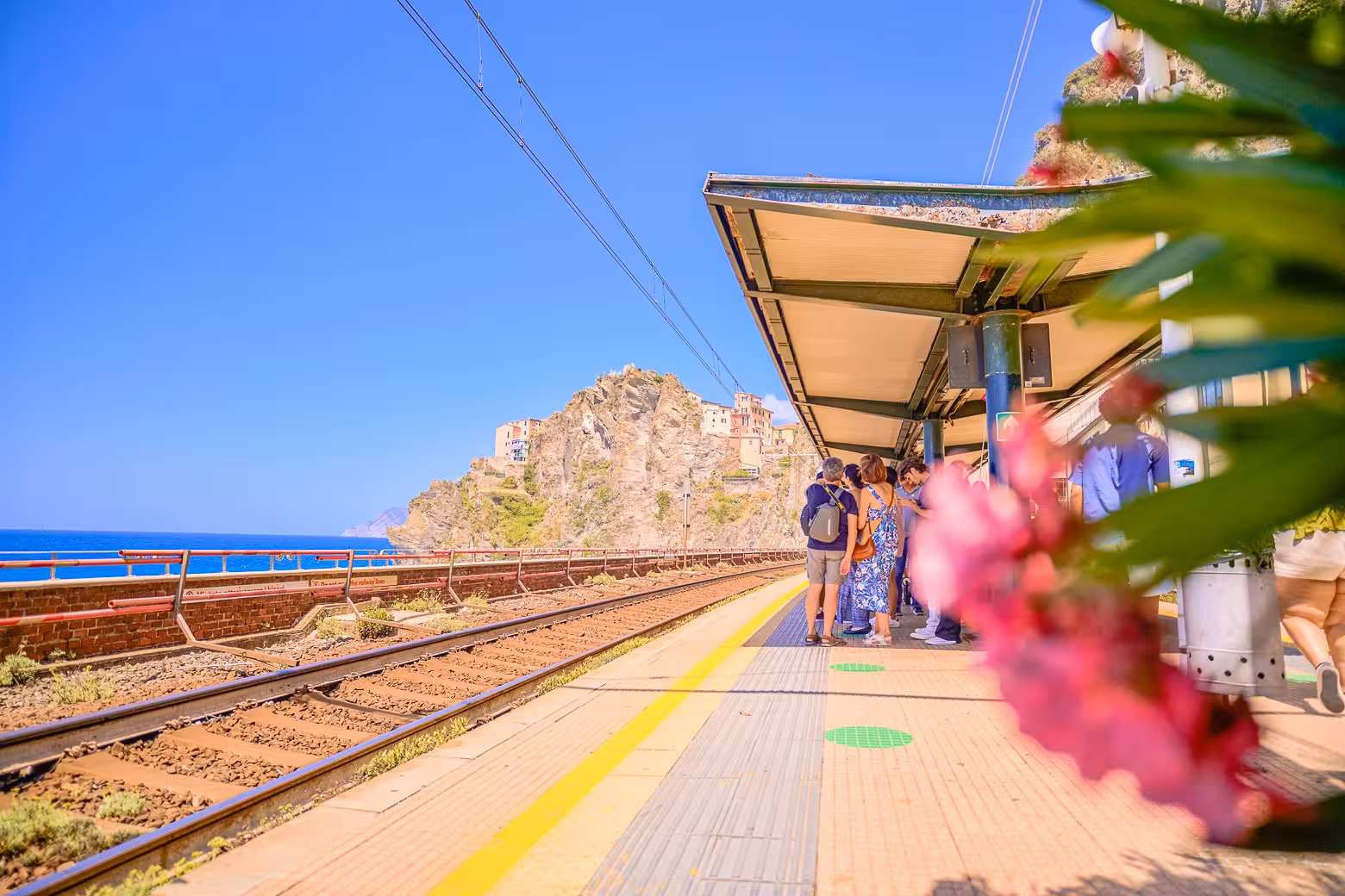Tourists waiting at a Cinque Terre train station with scenic coastal cliffs in the background.