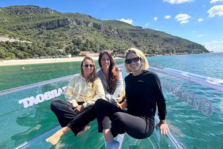 Three smiling people enjoy a sunny day on a clear boat, with stunning Sesimbra cliffs and beach in the background.