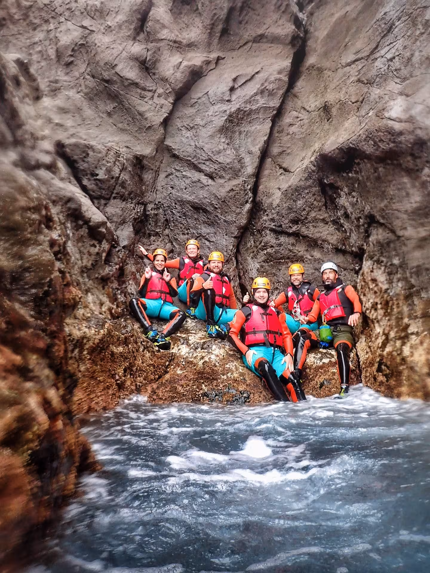 COASTEERING Freshman group in wetsuits and helmets resting on sea rocks as waves surge through a narrow cove