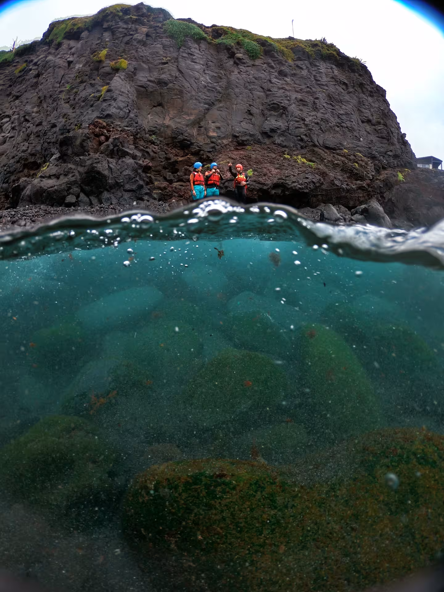 Coasteering Freshman participants in helmets and buoyancy aids by sea cliff, split-level ocean adventure view