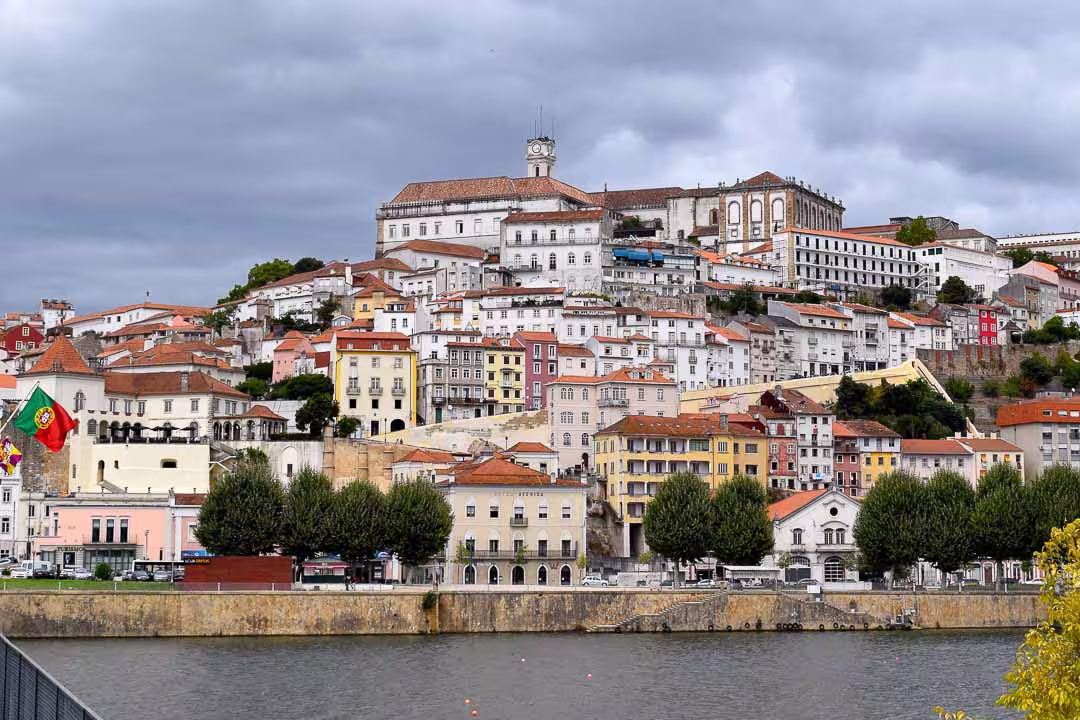 Scenic view of Coimbra's historic hillside architecture by the river, showcasing vibrant buildings under a cloudy sky.