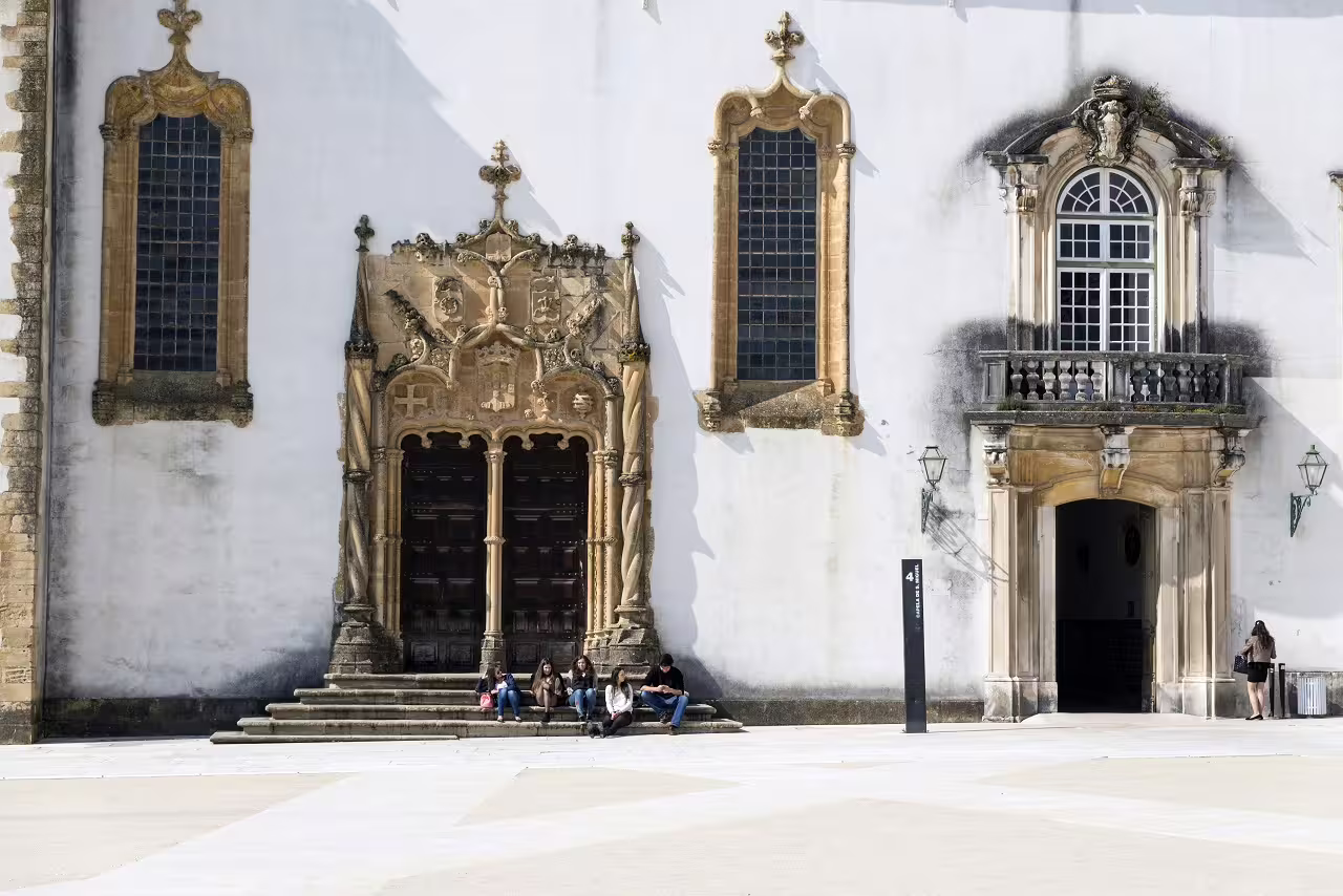 Historic facade of Coimbra University in Portugal with students sitting on steps, showcasing Gothic architecture and cultural heritage.