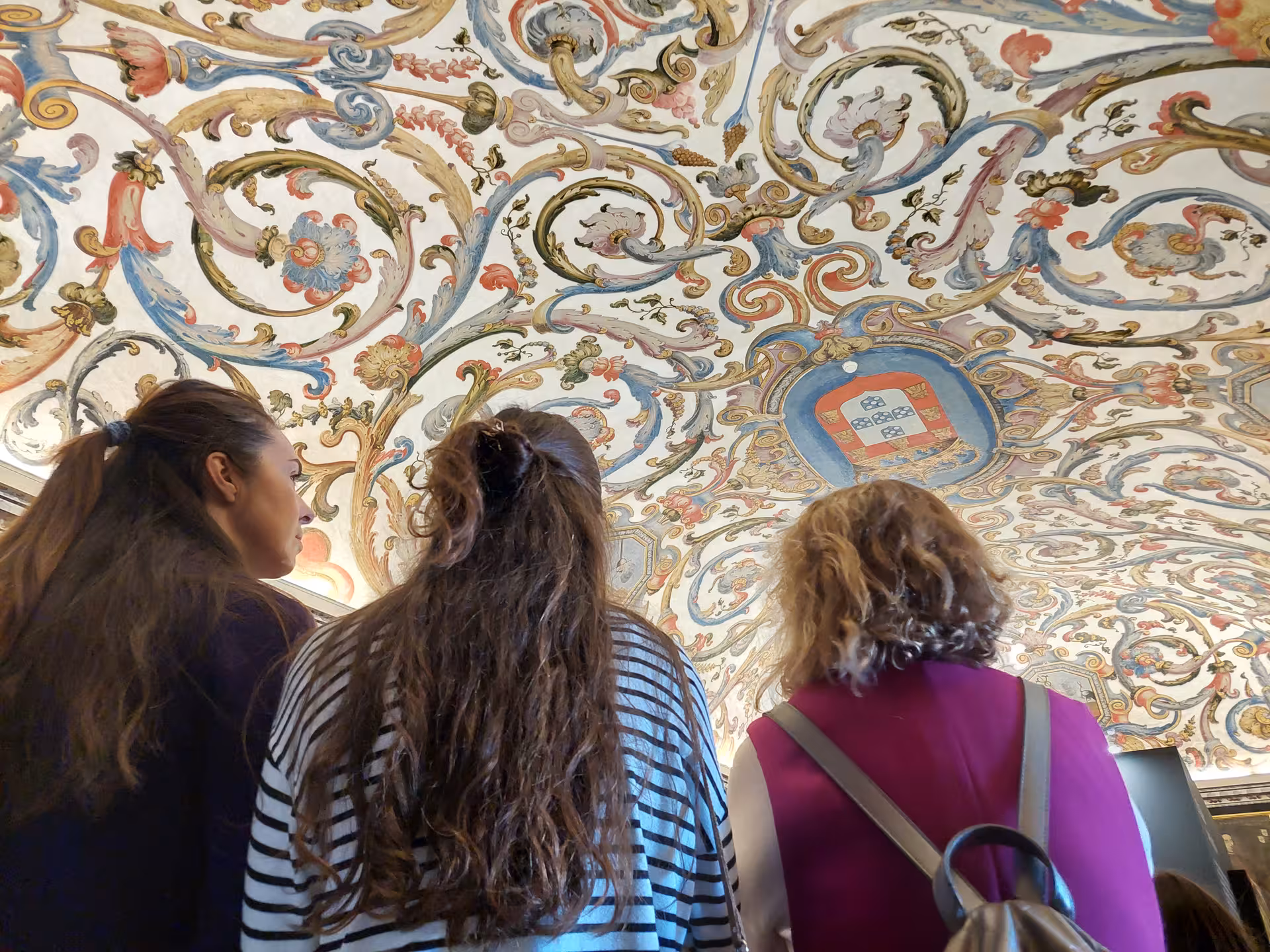 Visitors admire the intricate painted ceiling of Coimbra University's Joanina Library, showcasing rich historic details.