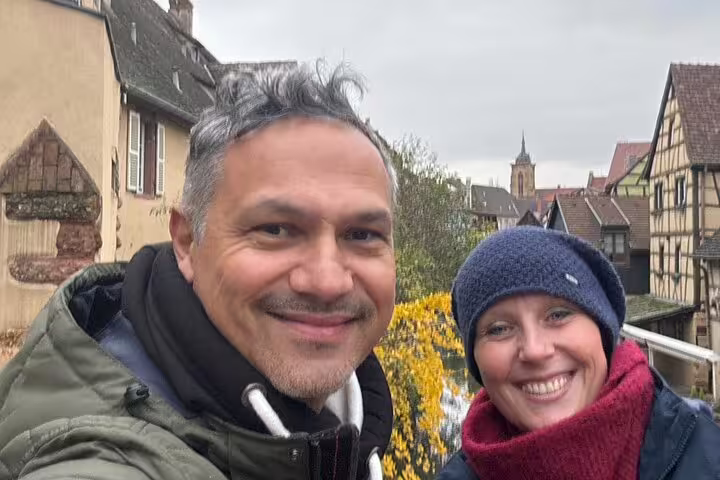 Smiling couple with Colmar old town rooftops and church tower, enjoying a self-guided scavenger hunt tour
