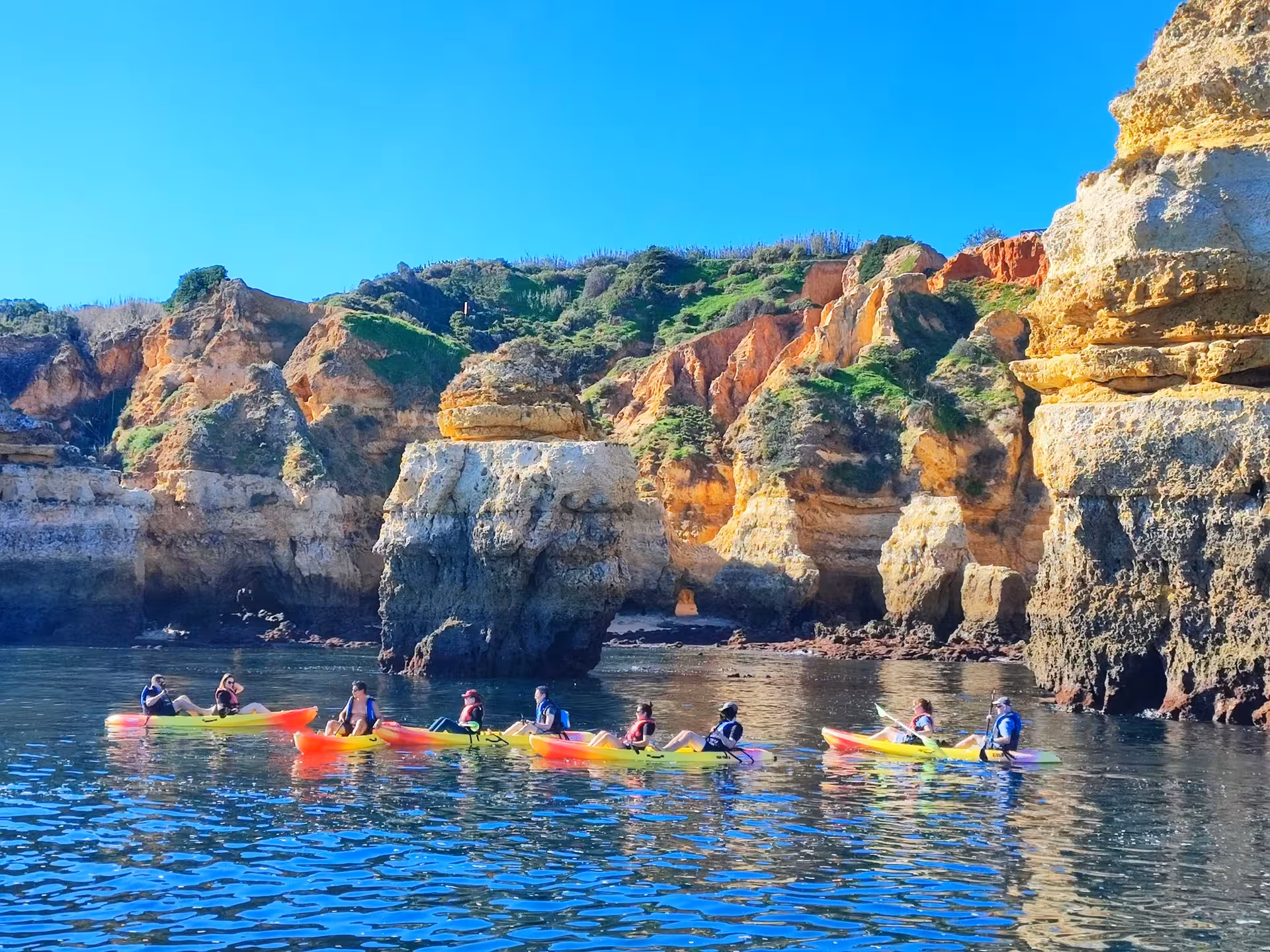 Colorful kayaks glide along the calm Lagos coastline, passing golden Algarve cliffs and sea stacks under clear blue skies