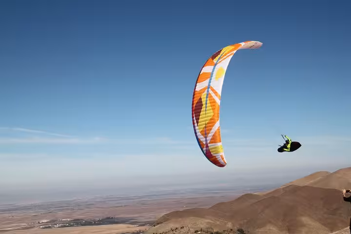 Colorful paraglider soaring over Agafay Desert hills under clear blue skies, showcasing Marrakech thrills.