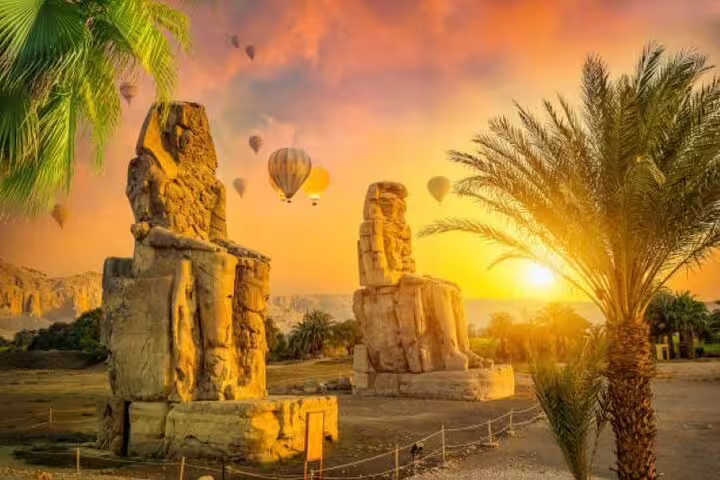 Colossi of Memnon at sunrise in Luxor with palm trees and hot air balloons, on Karnak and Valley of Kings tour