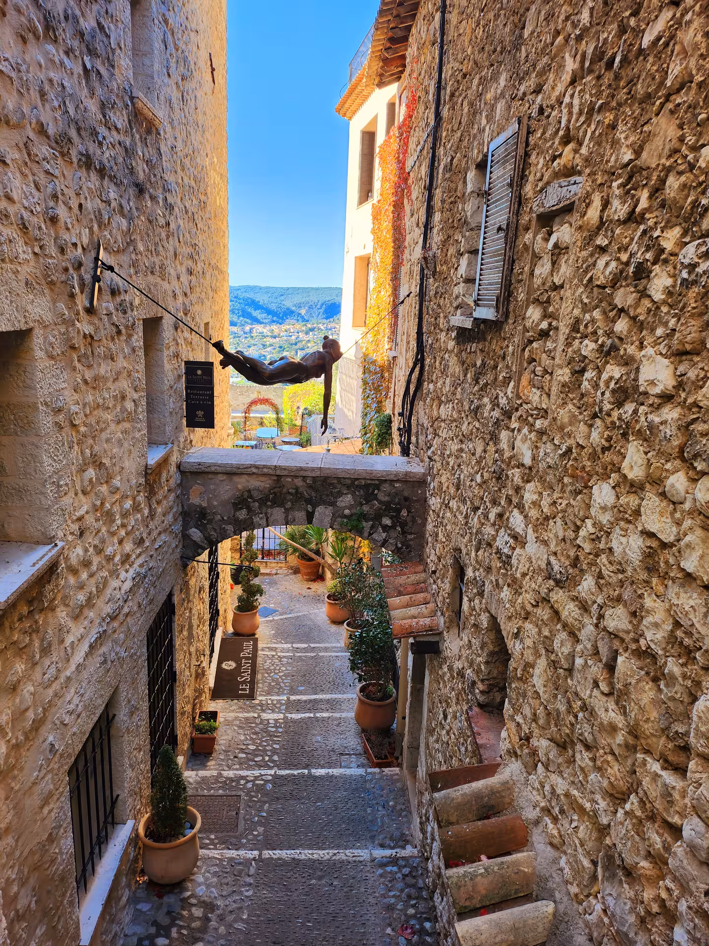 Charming cobblestone alley with rustic stone walls and a suspended sculpture, offering scenic countryside views.