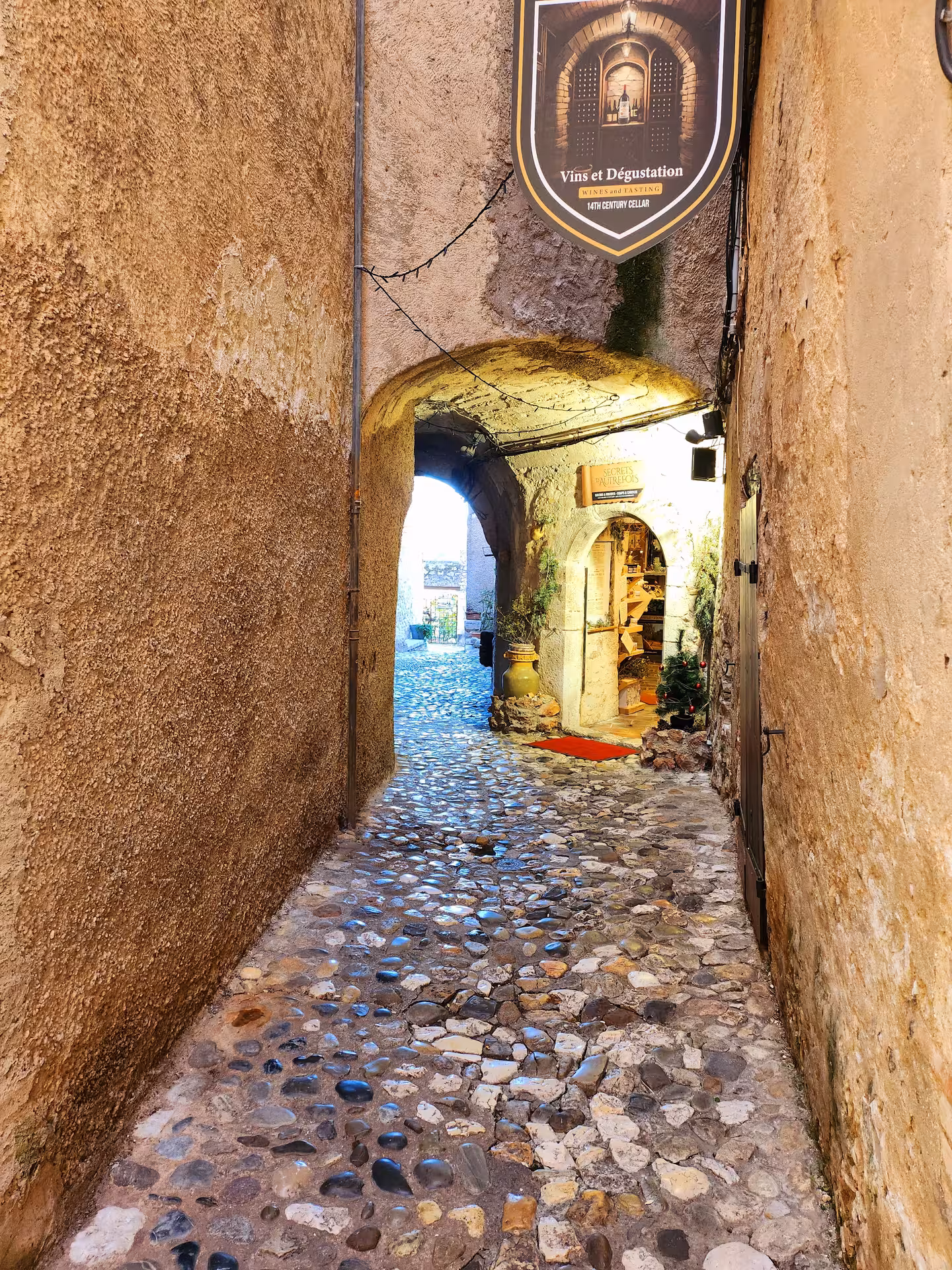 Cozy medieval alleyway with cobblestone path and inviting wine tasting sign, leading to a sunlit courtyard.