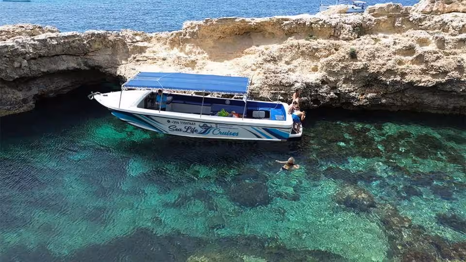 Boat in Comino Crystal Lagoon near sea cave, Malta, with swimmers in clear turquoise water on sunset tour