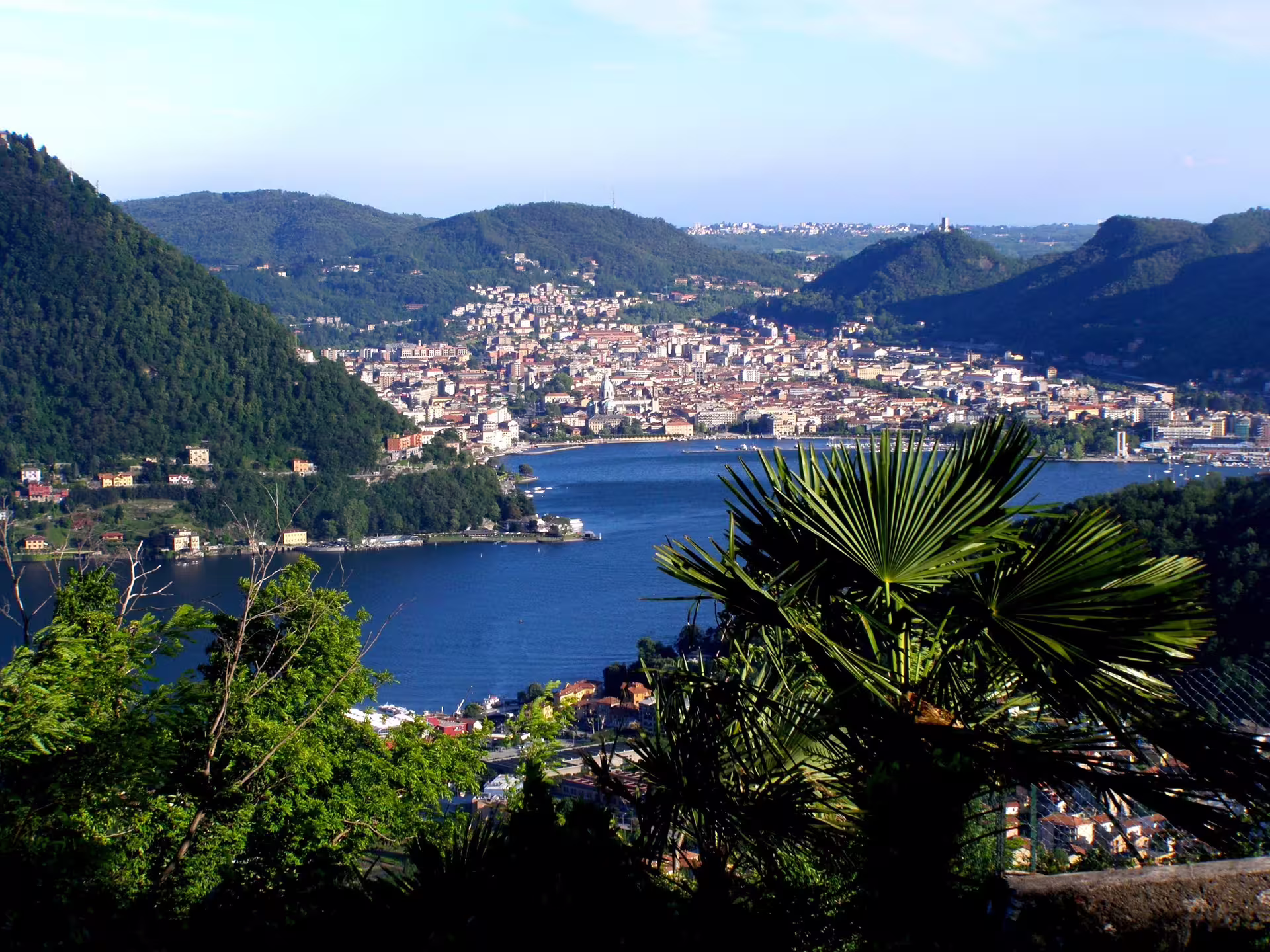 Panoramic view over Como city and deep blue Lake Como framed by lush green hills and palm trees in Italy