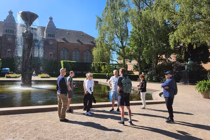 Visitors gather around a fountain in a lush Copenhagen garden during a guided walking tour on a sunny day.