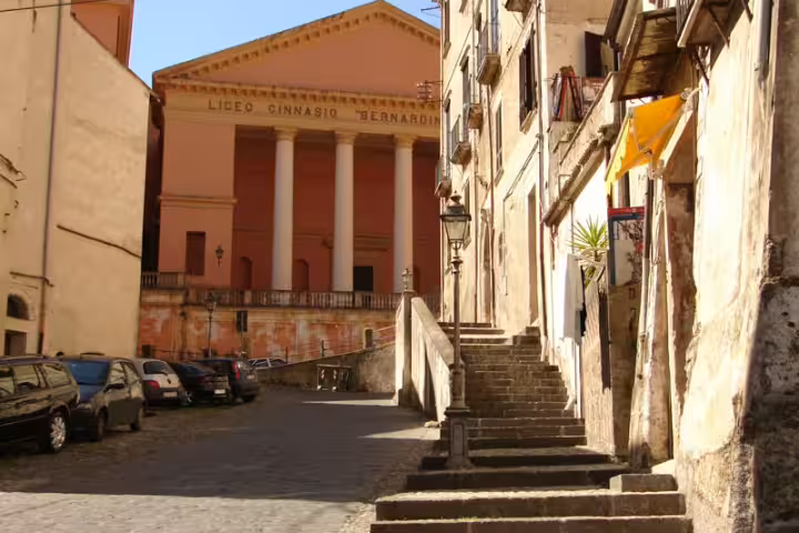 Sunlit old town street in Cosenza, Calabria leading to neoclassical Liceo Ginnasio building, ideal stop on wellness parks tour