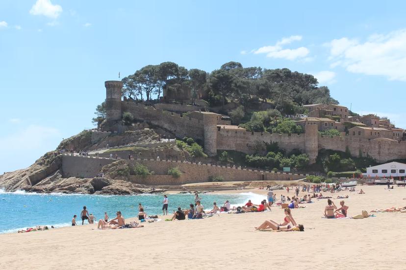 Sunbathers on a wide Costa Brava beach with Tossa de Mar fortress, day tour from Barcelona