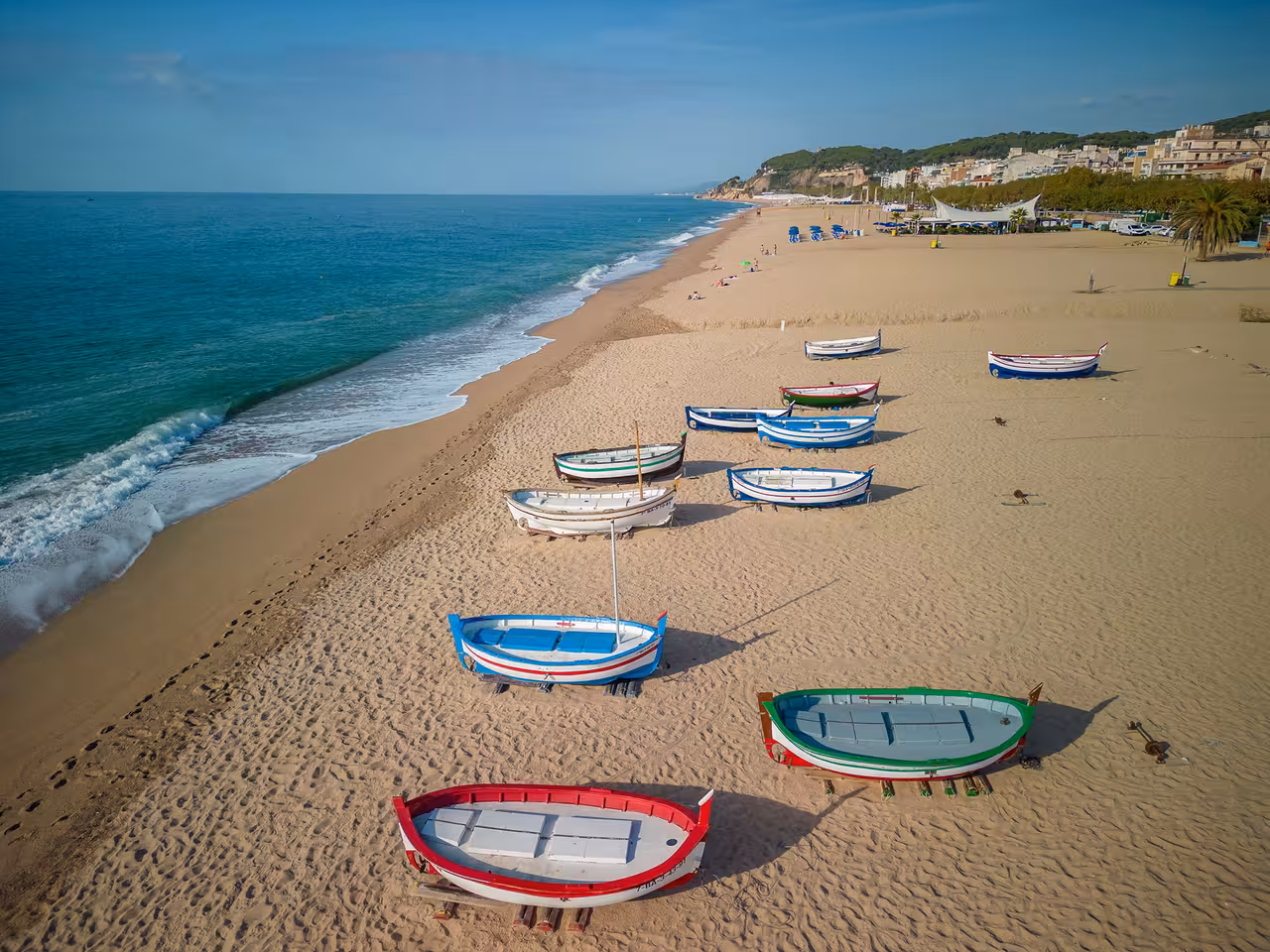 Colorful boats lined up on a sandy beach in Costa Brava with calm sea and clear blue skies.