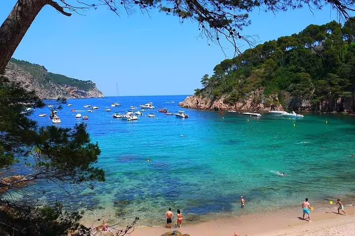 Vibrant Costa Brava beach with turquoise waters, boats, and sunbathers, perfect for a relaxing seaside tour.