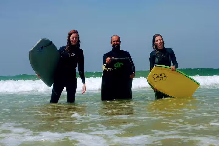 Three people in wetsuits holding bodyboards, standing in the ocean at Costa da Caparica, perfect for surf and yoga tours.
